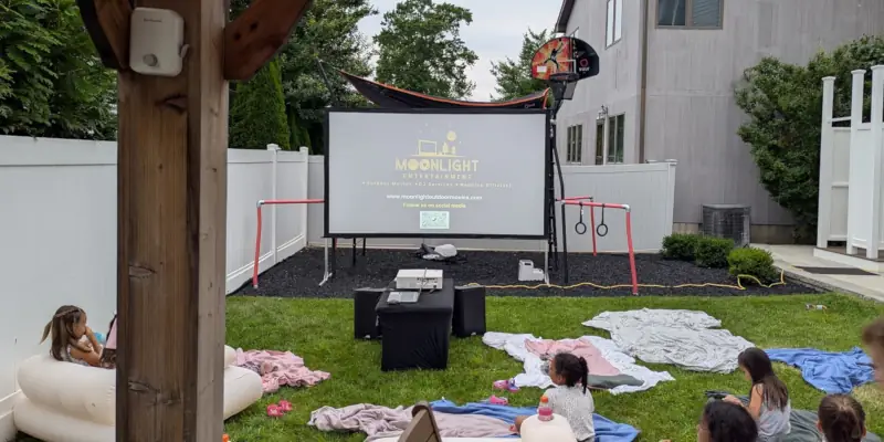 Backyard outdoor movie setup with a 10-foot screen displaying "Moonlight Entertainment" logo. Children sit on blankets and inflatable chairs, creating a cozy, playful atmosphere.