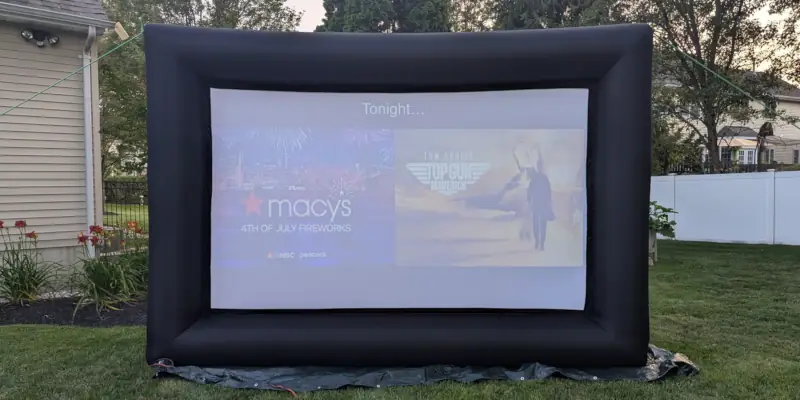 12-foot inflatable outdoor movie screen set up on a lawn, displaying "Macy's 4th of July Fireworks" and a "Top Gun: Maverick" preview, with trees and a house in the background.