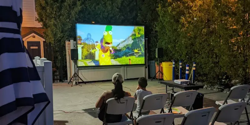 Outdoor movie screening at night with animated character on a 13-foot screen. Two people sitting in folding chairs watch attentively. Relaxed, cozy atmosphere.
