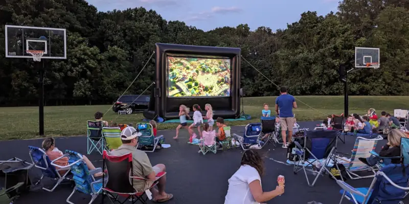 A group of people, including children, enjoy an outdoor movie night on foldable chairs facing a large 16-foot inflatable screen. The setting is relaxed and social.