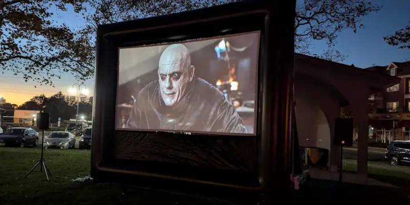 Outdoor movie screening at dusk, featuring a bald, pale character on a large 20-foot screen. Cars and trees in the background set a casual, eerie tone.
