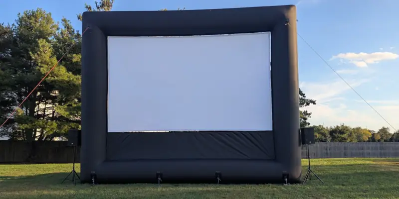 Inflatable 24-foot outdoor movie screen set up on a grassy field, surrounded by trees under a clear blue sky. The scene conveys a relaxed, open-air cinema atmosphere.
