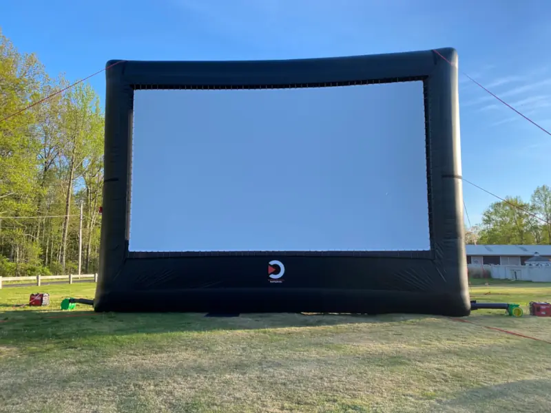 Large 30-foot inflatable outdoor movie screen set up on a grassy field, surrounded by trees and under a clear blue sky, creating a relaxed, open-air atmosphere.